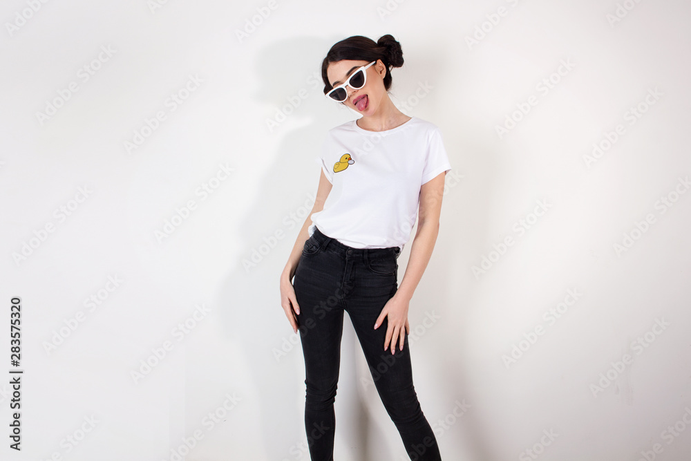 Fototapeta premium young funny brunette girl in a jeans and white shirt with sunglasses is standing and showing tongue on a white wall background, free space, fashion concept