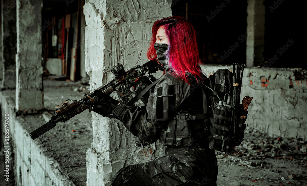 Red-hair airsoft woman in uniform with machine gun stand beside column. Soldier stand inside of broken building Red-hair airsoft woman in uniform with machine gun stand beside column. Soldier stand inside of broken building