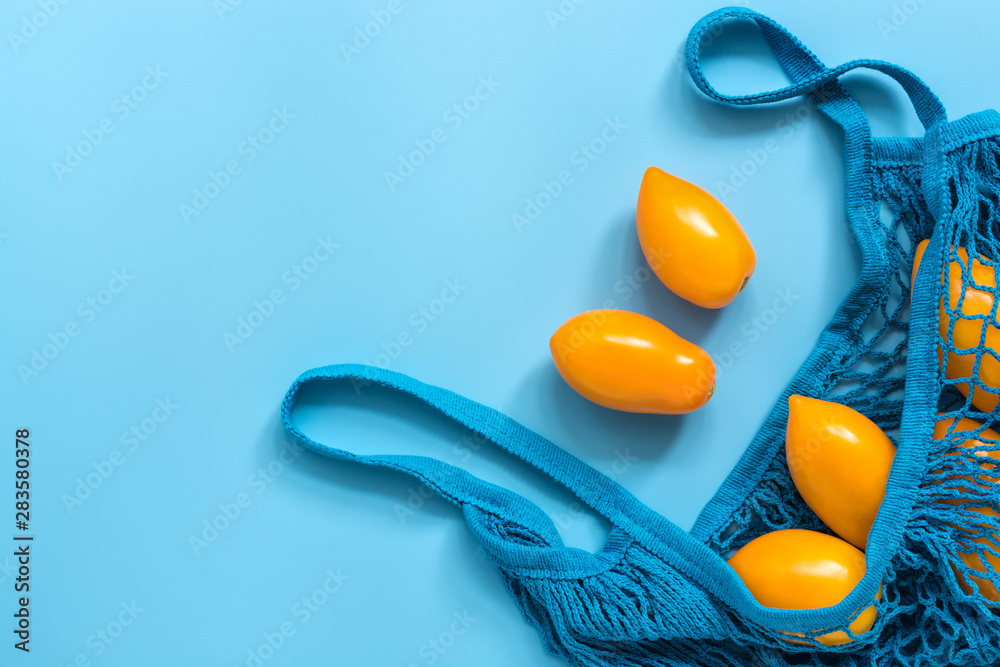 Fresh yellow tomatoes in a cotton string bag on a blue background. Flat lay, top view, copy space - obrazy, fototapety, plakaty 