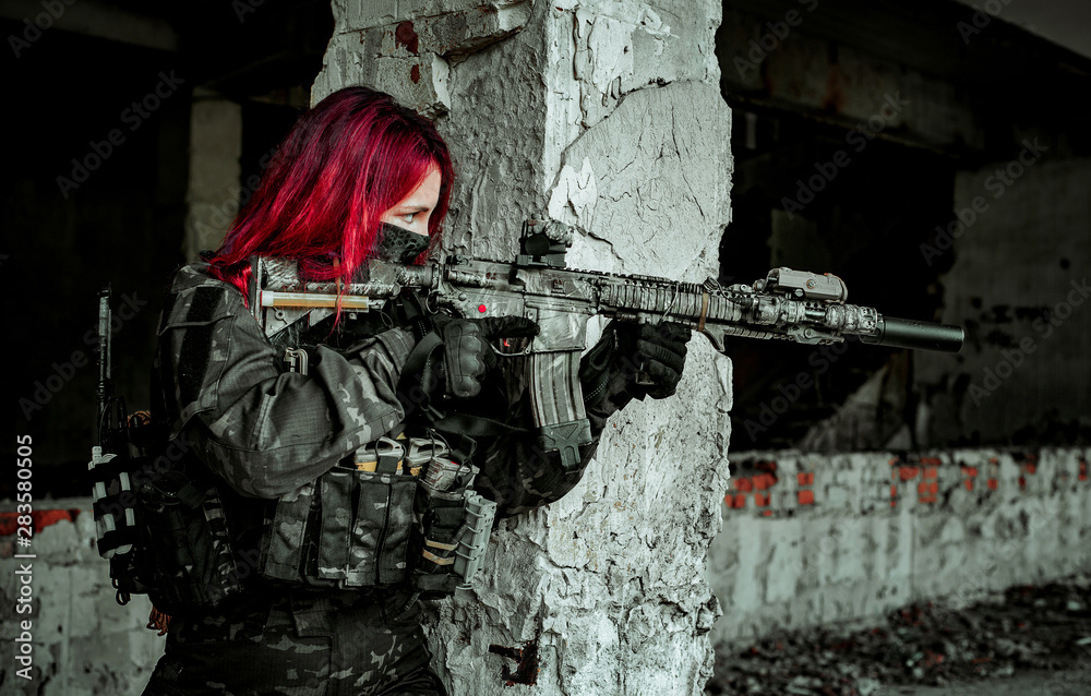 Airsoft red-hair woman in uniform, aim at the sight in machine gun. Close up soldier lurk beside concrete wall.