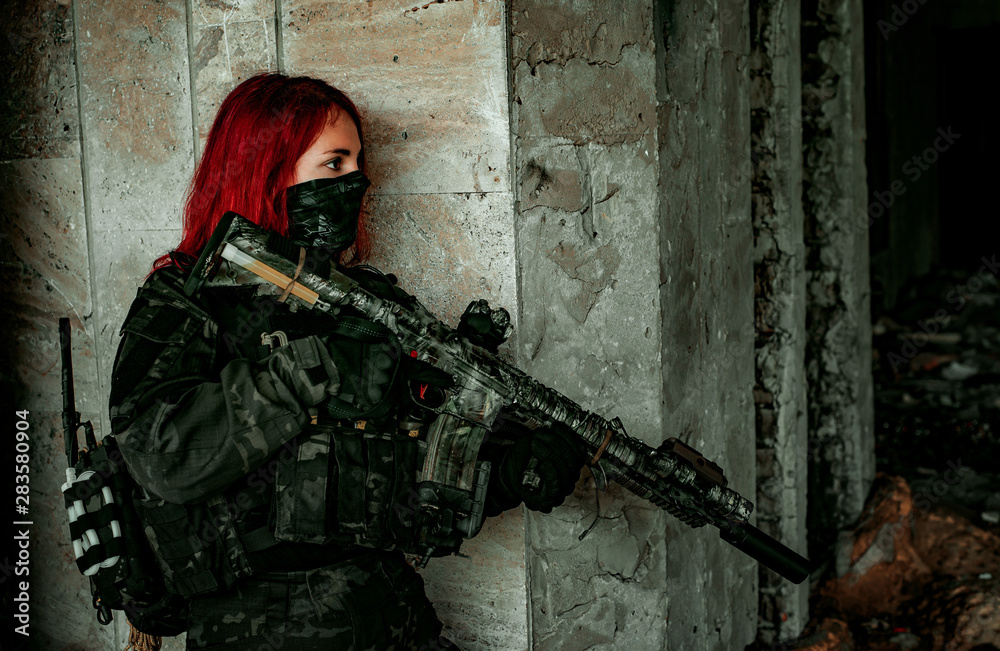 Fototapeta premium Airsoft red-hair woman in uniform with machine gun stand beside concrete wall. Soldier lurk inside broken building. Side view