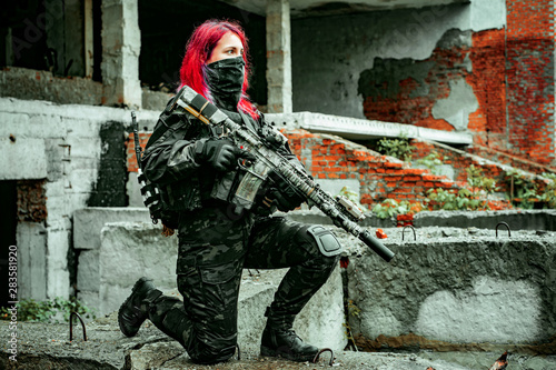Airsoft red-hair woman in uniform with machine gun standing on knee. Soldier on ruine. Side view
