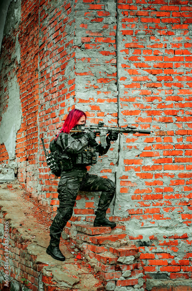 Fototapeta premium Airsoft red-hair woman in uniform with machine gun beside brick wall. Soldier aims at the sight on the ruins. Vertical photo side view