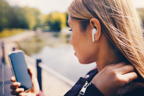 Young beautiful adult Caucasian girl walking outdoor in the park having chat communication with wireless headphones mobile reading talking listening using with her hand on sunset at summer