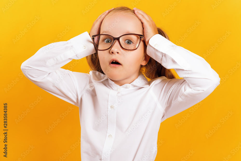 Portrait of a cheerful little girl in glasses on a yellow background. The child is shocked and holds his hands behind his head. Portrait of a cheerful little girl in glasses on a yellow background. The child is shocked and holds his hands behind his head.