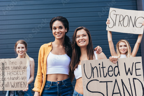 front view of smiling multiethnic feminists embracing and holding placard with inscription united we stand on street