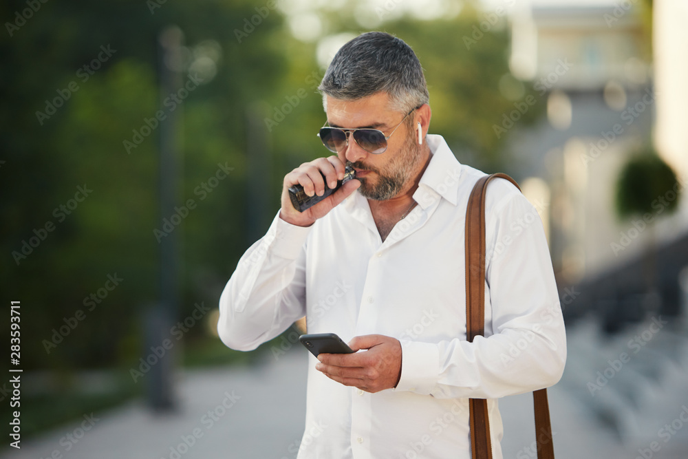 Middle-aged bearded thinking serious concentrated business man standing and smoking electronic cigarette. Blows smoke through the mouth. In-ear wireless earphone. Office centre background Middle-aged bearded thinking serious concentrated business man standing and smoking electronic cigarette. Blows smoke through the mouth. In-ear wireless earphone. Office centre background
