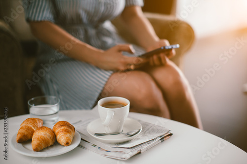 Cropped photo of woman in cafe drinking coffee, eating croissant and using mobile phone