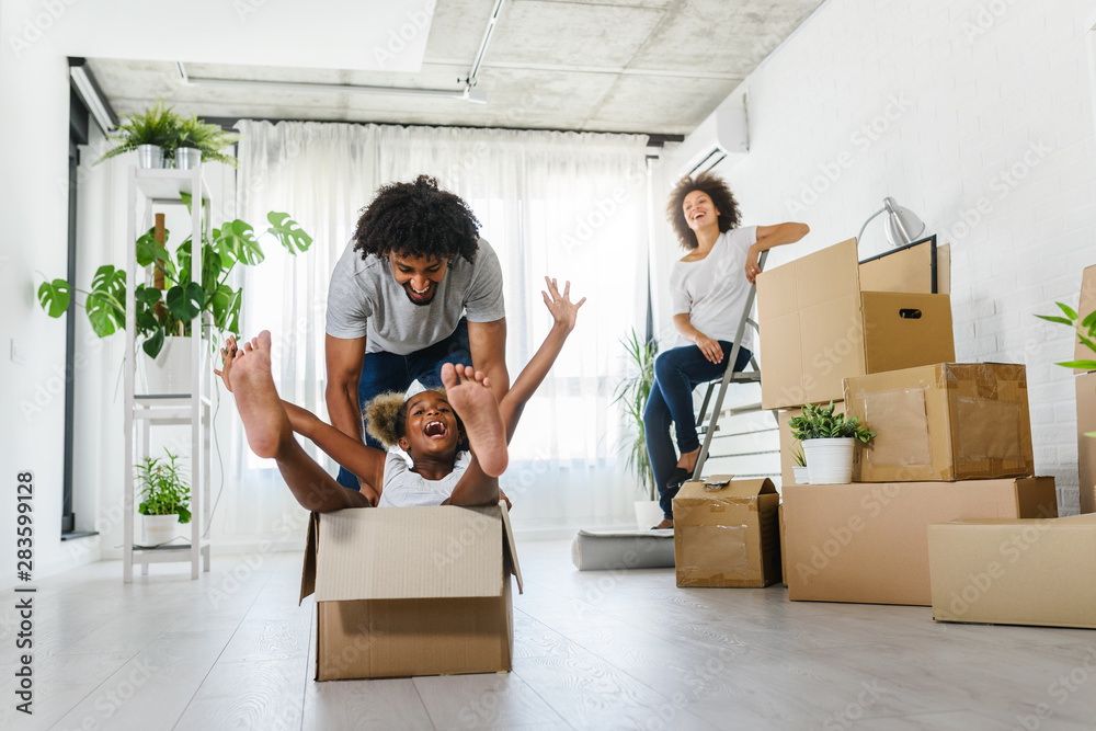 Happy playful  African American family moving in new apartment, little preschooler daughter sitting in cardboard boxes, father rolling her,  purchase property concept - obrazy, fototapety, plakaty 