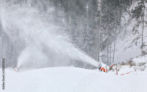 Orange and blue snow making cannon spreading ice crystals over ski piste, trees in background