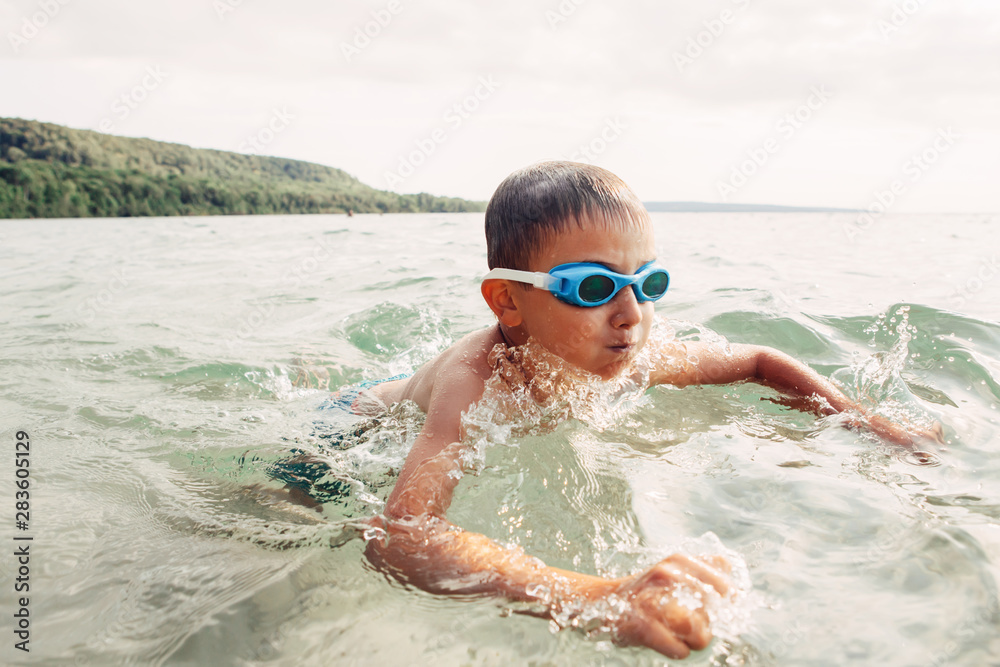 Cute funny Caucasian boy swimming in lake river with underwater goggles. Child diving in water on beach. Authentic real lifestyle happy childhood. Summer fun outdoor aquatic activity.