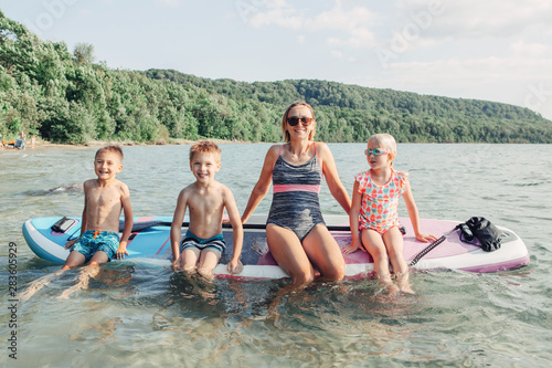 Caucasian woman parent sitting on paddle sup surfboard in water with kids children. Modern outdoor family activity. Individual summer aquatic recreation sport hobby. Healthy lifestyle.