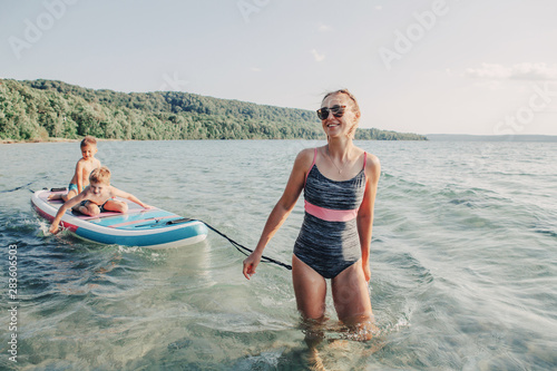 Caucasian woman parent riding kids children boys on paddle sup surfboard in water. Modern outdoor summer fun family activity. Individual aquatic recreation sport hobby. Healthy lifestyle. photo