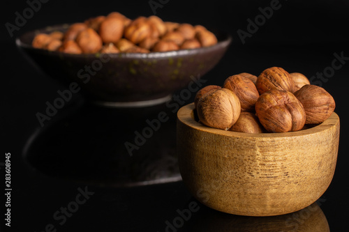 Lot of whole ripe brown hazelnut in dark ceramic bowl in tiny wooden bowl isolated on black glass