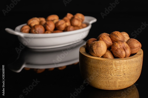 Lot of whole ripe brown hazelnut in white oval ceramic bowl in tiny wooden bowl isolated on black glass