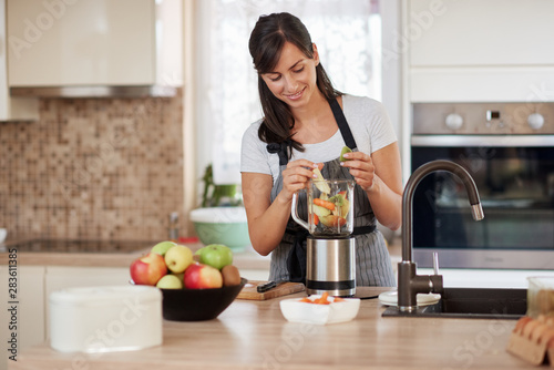 Beautiful Caucasian smiling woman in apron putting vegetables and fruits in blender while standing in kitchen.