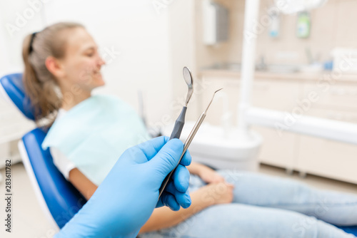 Close-up hand of dentist in the glove holds tool probe and mirror. The patient in the dental chair at the background. Dental work in clinic. Office where dentist conducts inspection and concludes.