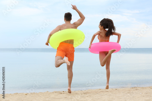 Happy young couple having fun with inflatable rings on beach near sea