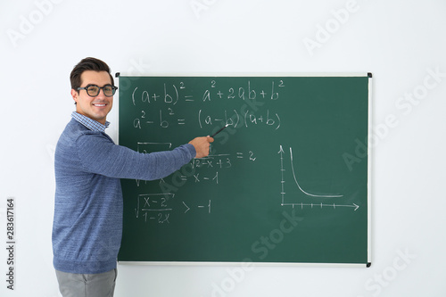 Young teacher explaining math formulas written on chalkboard in classroom