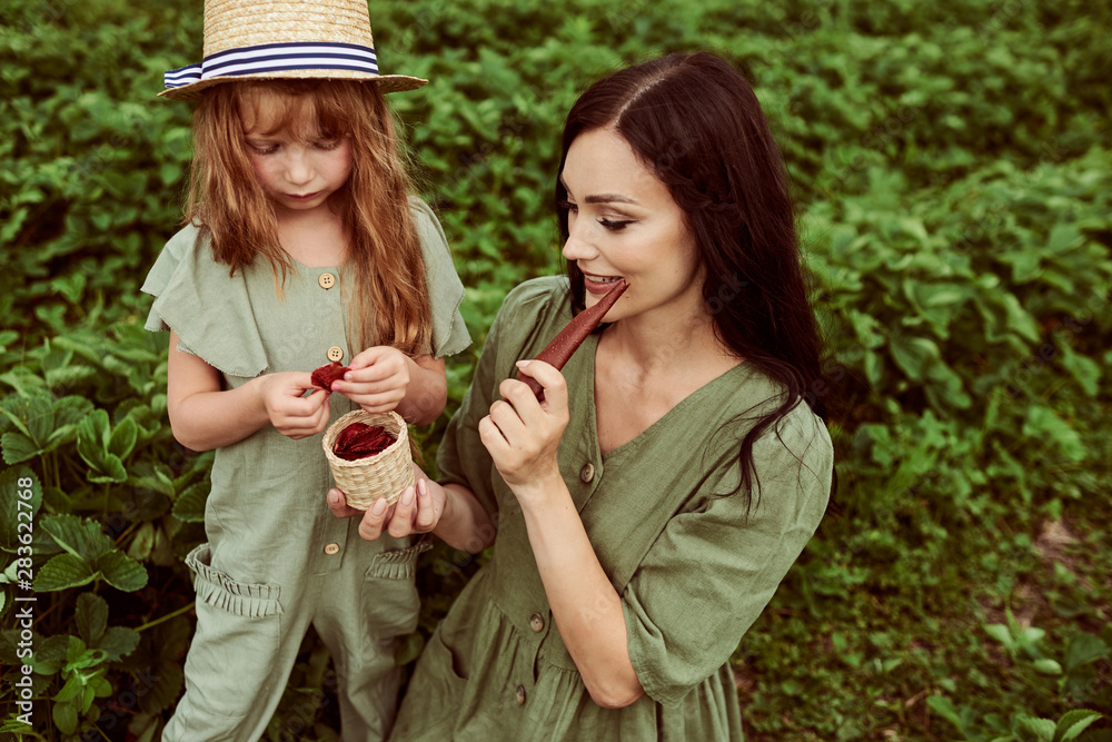 Fototapeta premium Beautiful young mother with her daughter having fun on a green field with strawberries
