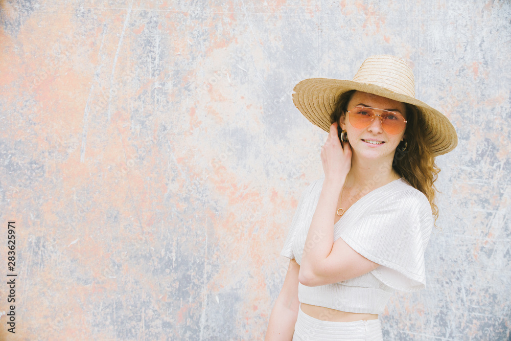 Millenial trendy Hipster teenager enjoys and relaxes during a walk in city center, she has a straw hat and orange lens sunglasses and white minimalistic dress, portraiture series,
