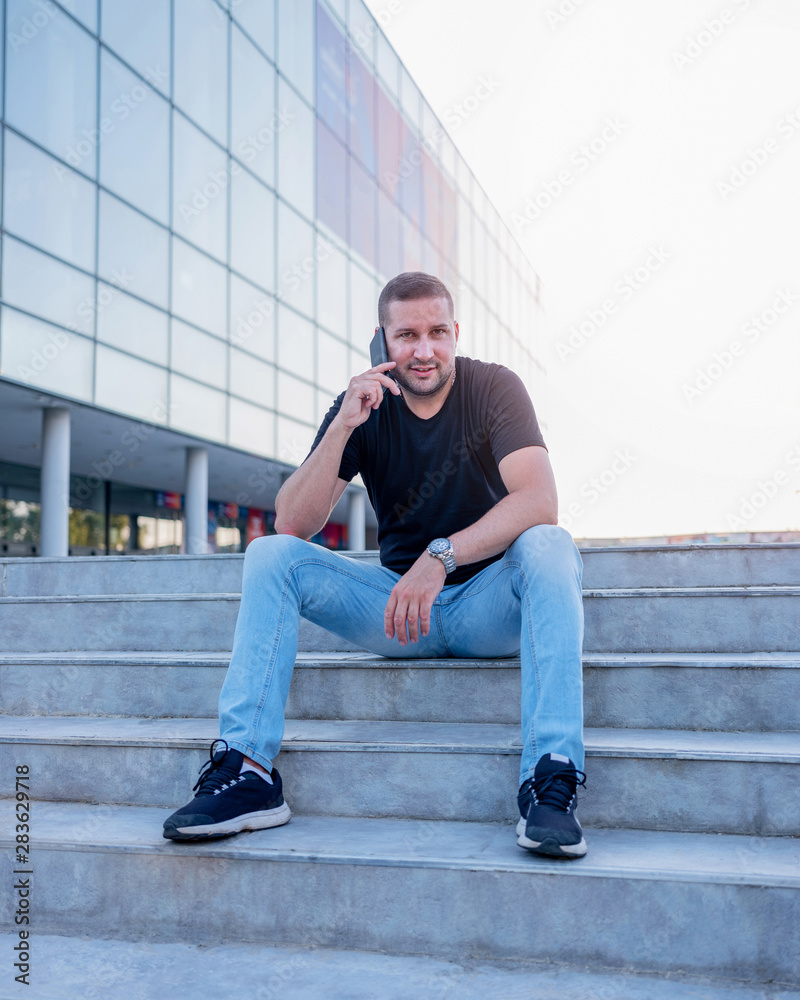 A handsome guy sitting on the stairs and talking on his smartphone.