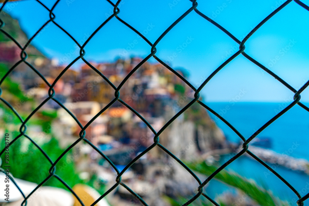 The famous village of Manarola behind the iron fence on the hill. A beautiful blue sky and sunny day of the European summer in Liguria, Italy. The famous village of Manarola behind the iron fence on the hill. A beautiful blue sky and sunny day of the European summer in Liguria, Italy.