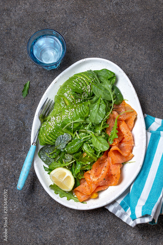 Low carbs salad. Spinach, rucola salad with avocado and salmon. Black concrete background, white plate, top view photo