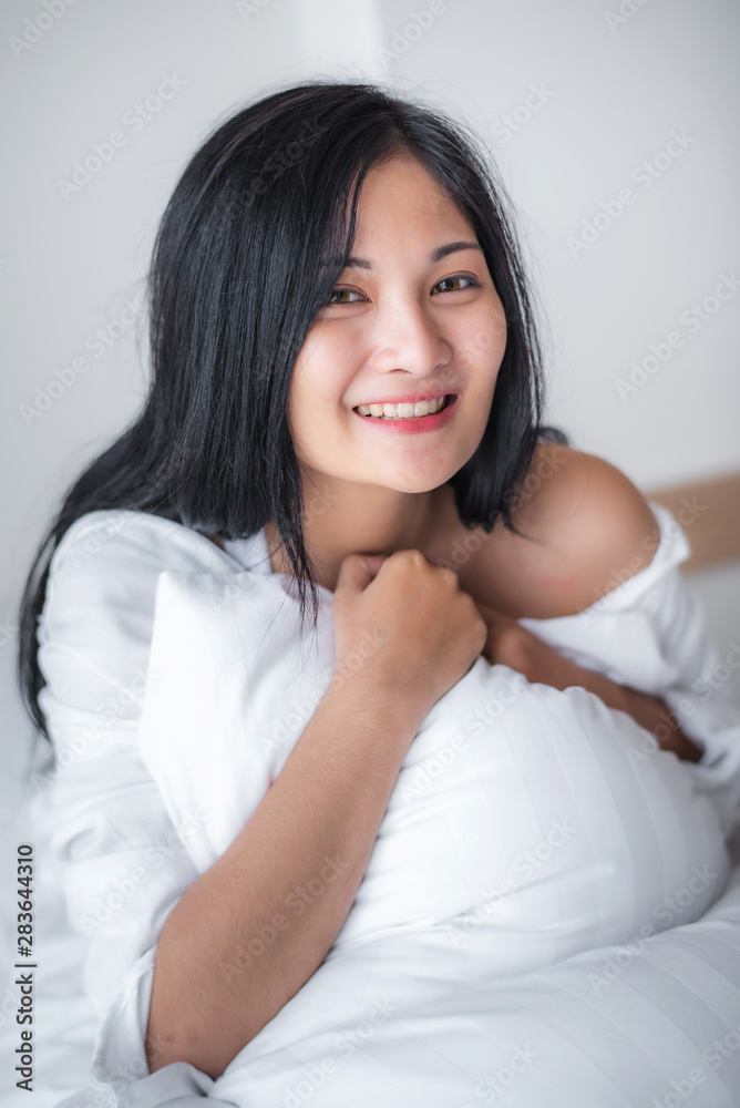 Beautiful young pretty Asian woman wake up and make happy smile with white shirt at the white bed in the morning. Beautiful young pretty Asian woman wake up and make happy smile with white shirt at the white bed in the morning.