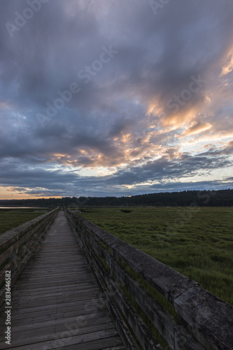 wooden boardwalk streching far across a lush green wetlands below a bright cloudy sunset