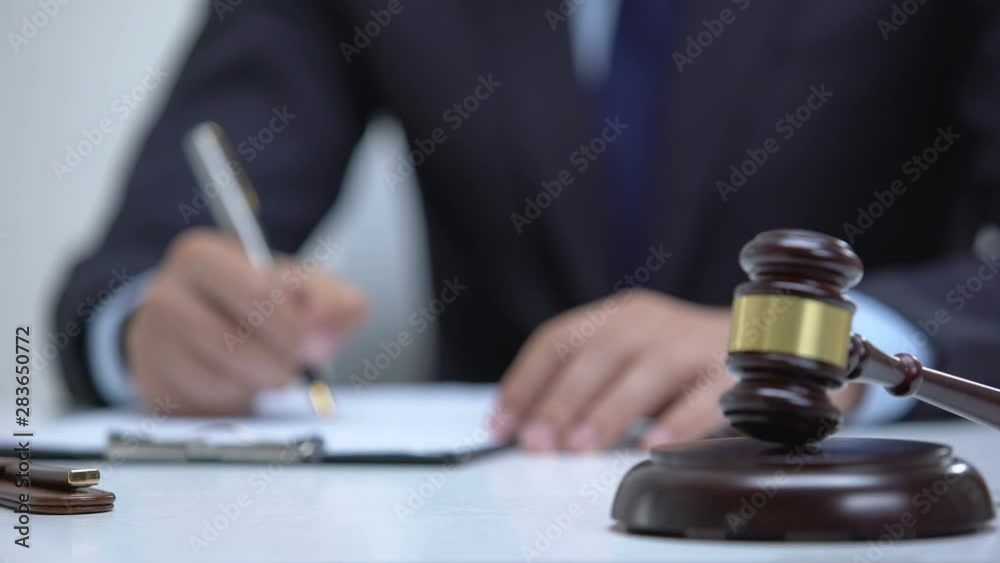 Gavel on block as jurisdictional symbol, attorney signing evidence on background Gavel on block as jurisdictional symbol, attorney signing evidence on background