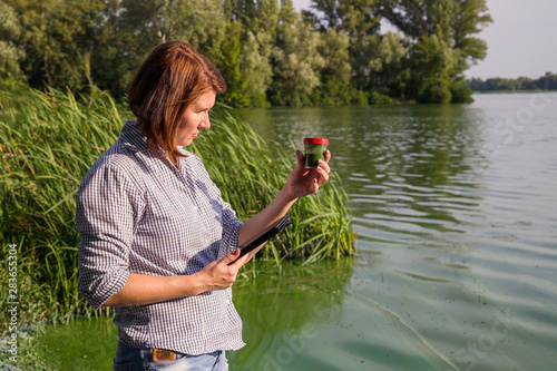 Tableau sur toile woman ecologist on river bank examines sample of green algae and holds tablet