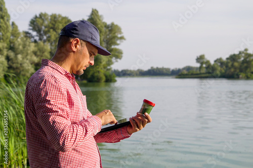 side view of ecologist on river bank entering data on green algae on tablet