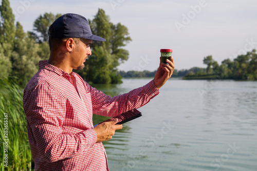Fotografia ecologist on river bank examines container with green algae and holds tablet