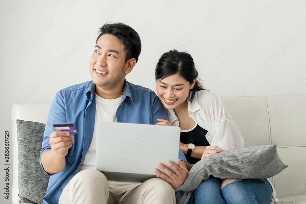 Happy Asian couple shopping online with holding credit card and laptop and sitting at home together, lifestyle