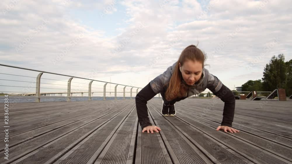 Panning of young sporty Caucasian woman doing push-ups on wooden embankment near water reservoir Panning of young sporty Caucasian woman doing push-ups on wooden embankment near water reservoir