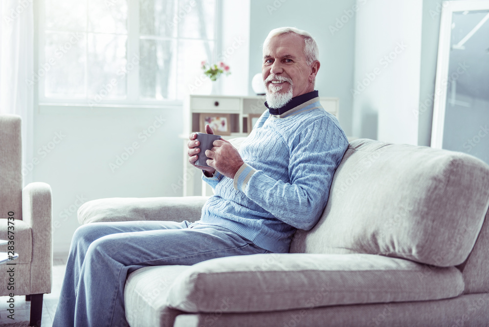 Beaming retired man wearing jeans and blue sweater drinking some tea