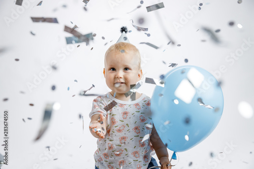 A little cute happy girl rejoices and laughs on her first birthday on a white background. Slapstick with confetti, a balloon filled with helium, a silver cap.