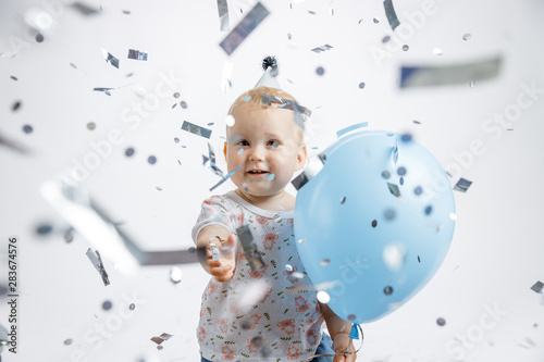 A little cute happy girl rejoices and laughs on her first birthday on a white background. Slapstick with confetti, a balloon filled with helium, a silver cap.