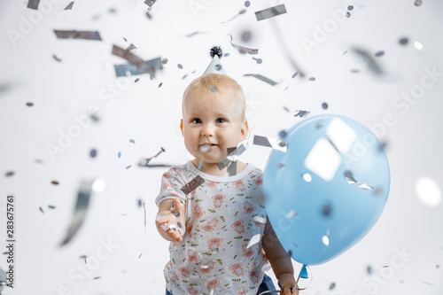 A little cute happy girl rejoices and laughs on her first birthday on a white background. Slapstick with confetti, a balloon filled with helium, a silver cap.