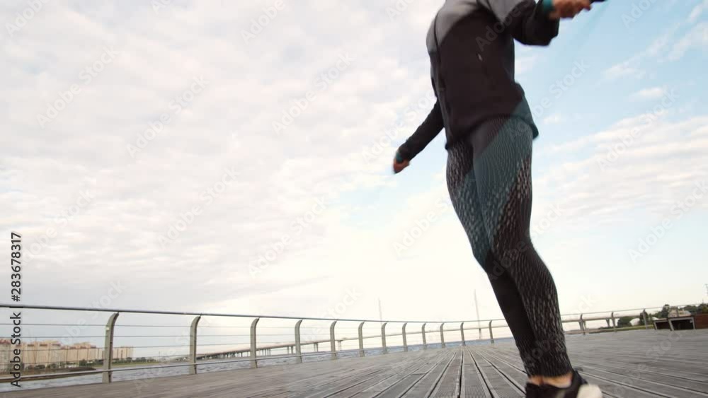 Tilt down of young sporty Caucasian brunette jumping using jump rope on wooden embankment outdoors while listening to music Tilt down of young sporty Caucasian brunette jumping using jump rope on wooden embankment outdoors while listening to music