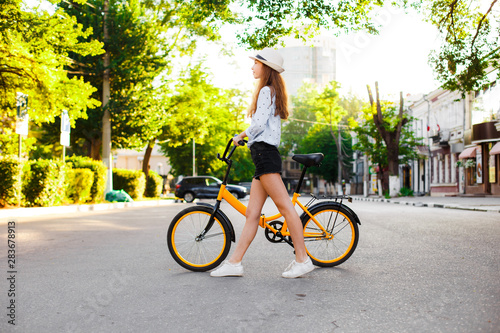 Girl teenager in a hat with a yellow bike walks through the summer city.
