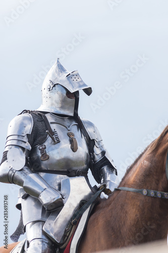 A man knight with half opened helmet riding a brown horse holding a sword