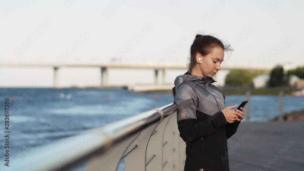 Medium shot of young Caucasian woman standing outdoors on embankment with wireless headphones in her ears and smartphone in her hands Medium shot of young Caucasian woman standing outdoors on embankment with wireless headphones in her ears and smartphone in her hands
