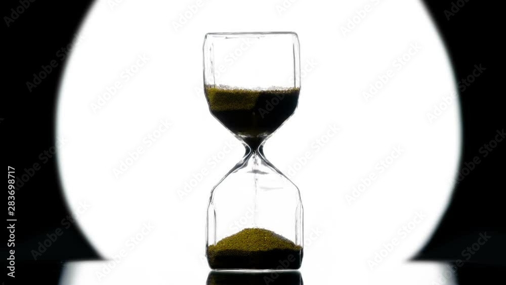 Sandy Watches with Gold Sand. Hourglass in Style Noir.Golden sand is strewed against the background of a bright round beam of a spotlight on a black. Filmed at a speed of 120fps Sandy Watches with Gold Sand. Hourglass in Style Noir.Golden sand is strewed against the background of a bright round beam of a spotlight on a black. Filmed at a speed of 120fps