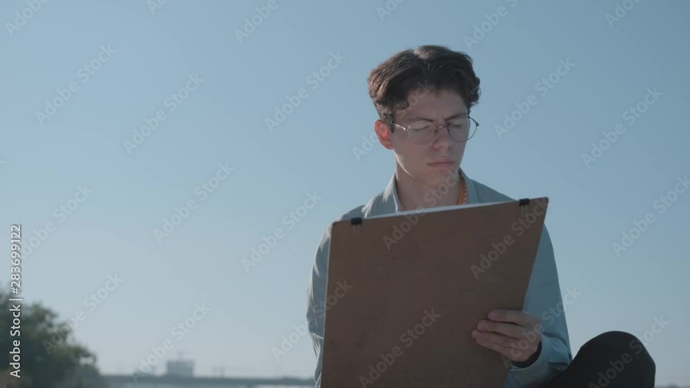 Pensive young caucasian man in striped t-shirt and denim shirt draws on the air. Taken from below. Portrait of modern hipster painter in glasses. He is draws with pencil and looks around. Pensive young caucasian man in striped t-shirt and denim shirt draws on the air. Taken from below. Portrait of modern hipster painter in glasses. He is draws with pencil and looks around.