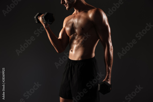 Concentrated handsome young strong sportsman posing isolated over black wall background holding dumbbells.