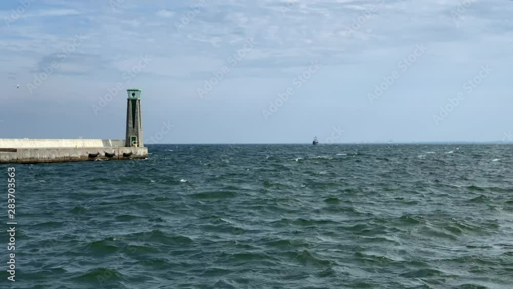 Slowmotion of a green sea waves crashing into concrete pier with lighthouse at the end in a bright day. Waves hitting breakwater. Slowmotion of a green sea waves crashing into concrete pier with lighthouse at the end in a bright day. Waves hitting breakwater.