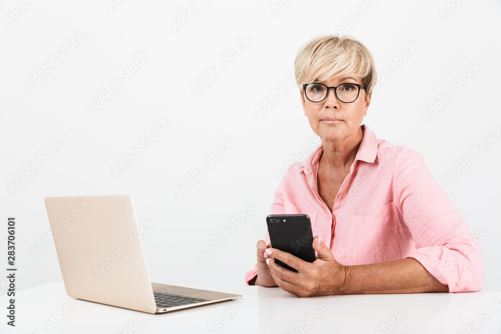 Portrait of charming middle-aged woman wearing eyeglasses holding cellphone and using laptop computer Portrait of charming middle-aged woman wearing eyeglasses holding cellphone and using laptop computer