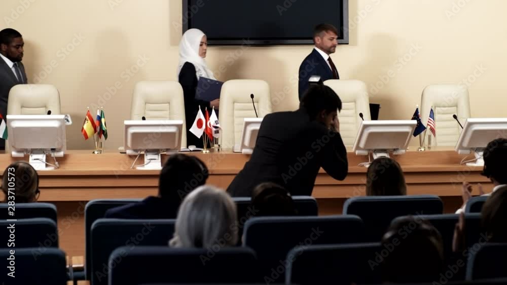 Multiethnic participants of political press conference walking into meeting room and taking their seats while photographer taking pictures and audience clapping Multiethnic participants of political press conference walking into meeting room and taking their seats while photographer taking pictures and audience clapping